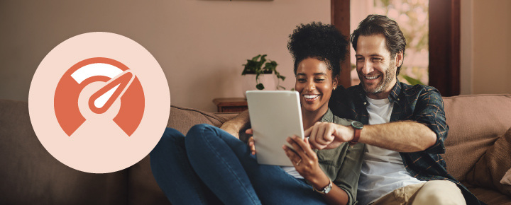 A cozy, indoor scene where a smiling man and woman are sitting on a beige couch looking at a tablet.