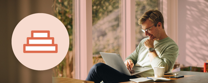 Man working on a laptop at a table, with a coffee cup, notebook, and pen, against a background of a large window and greenery.