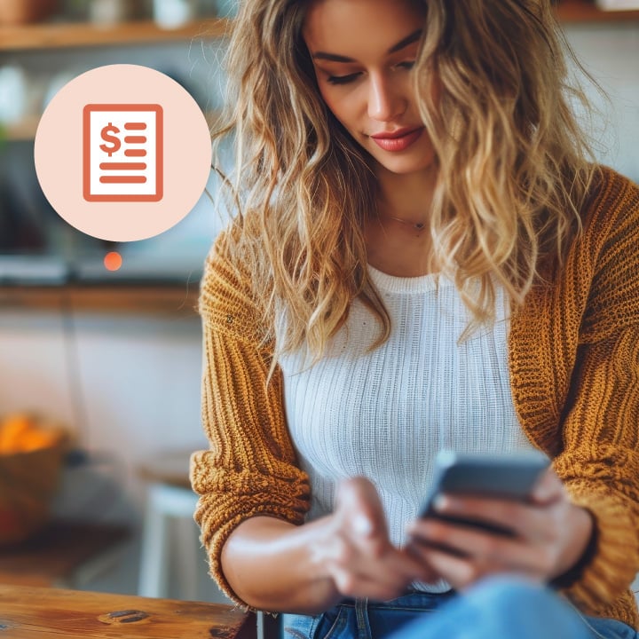 Young woman sitting indoors, looking at her smartphone, with a finance-related icon in the upper left corner.