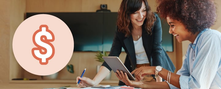 Two women smiling and interacting with a tablet in an office, with a dollar sign icon nearby.