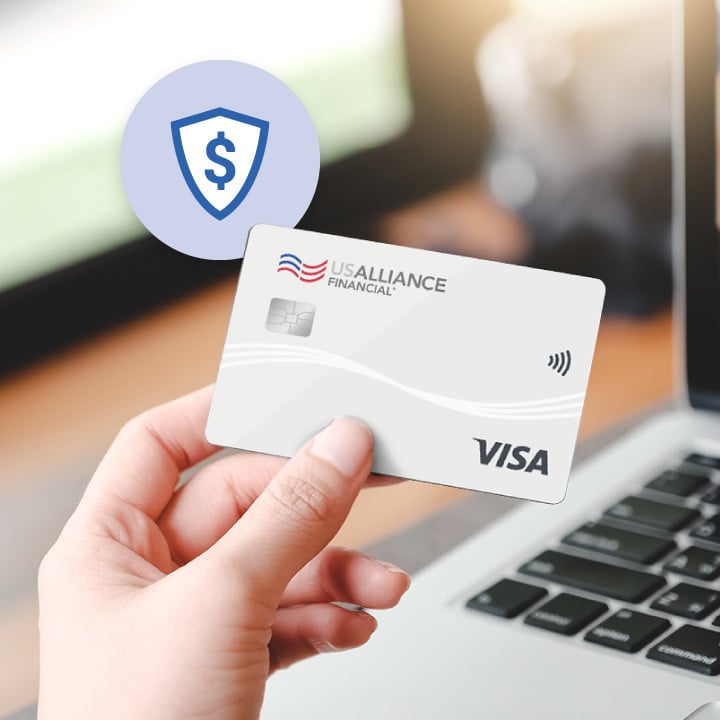 Hand holding a USALLIANCE Financial VISA Secured credit card in front of a laptop.