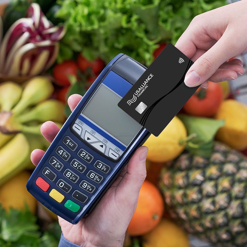 A person holding a POS terminal with another person holding a contactless card near it, set against a background of assorted fresh produce.