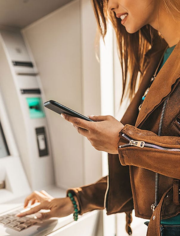 Woman in leather jacket using an ATM while holding a smartphone.